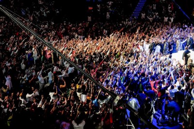 Un chanteur sur scène, vêtu de blanc, levant un bras sous des lumières de spectacle et un écran en arrière-plan.