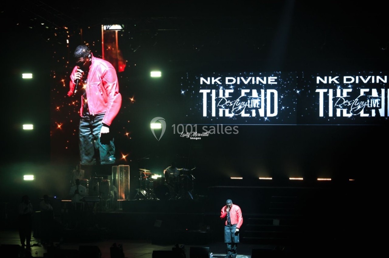 Un artiste chante sur scène dans une salle sombre, éclairée par des projecteurs et des écrans affichant son nom.