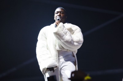 Un chanteur sur scène, vêtu de blanc, levant un bras sous des lumières de spectacle et un écran en arrière-plan.