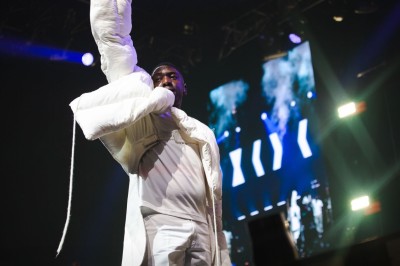 Un chanteur sur scène, vêtu de blanc, levant un bras sous des lumières de spectacle et un écran en arrière-plan.