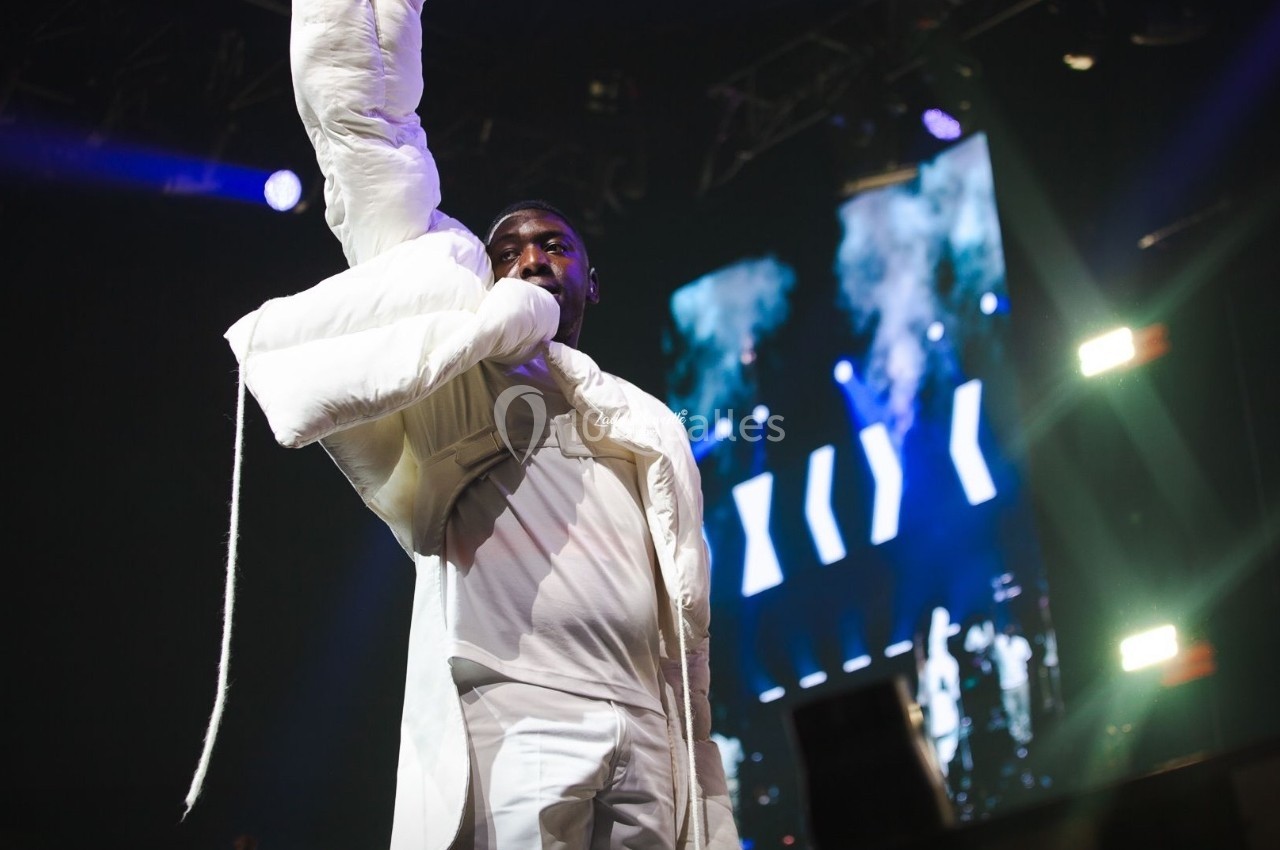 Un chanteur sur scène, vêtu de blanc, levant un bras sous des lumières de spectacle et un écran en arrière-plan.