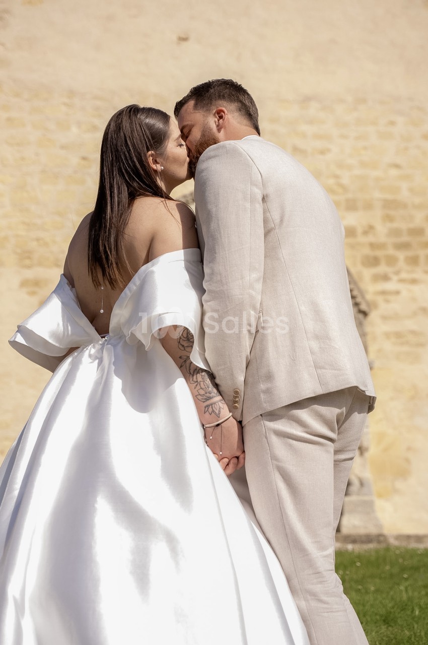 Des mariés en tenue claire s'embrassent en extérieur devant un mur de pierre ocre, la mariée de dos dans une robe blanche à large jupe satinée.