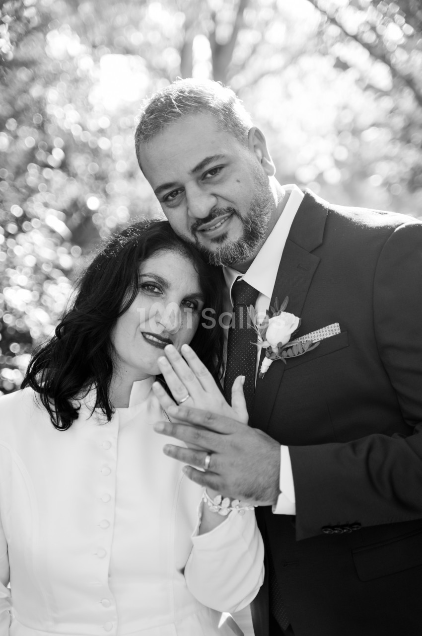 Couple de mariés en noir et blanc, la femme en robe blanche boutonnée appuyée contre l'homme en costume sombre à fleur à la boutonnière, devant un fond de feuillage flou.