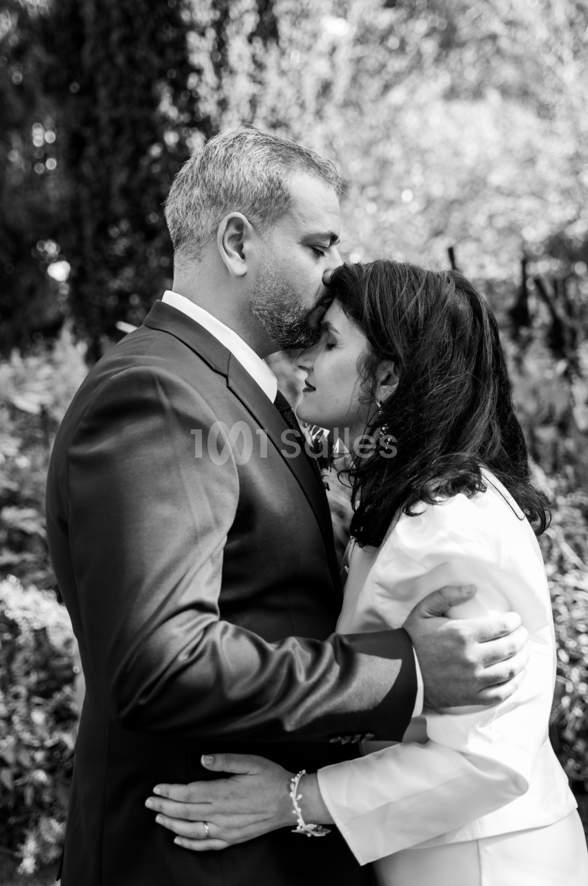 Photo en noir et blanc d'un homme en costume sombre embrassant le front d'une femme aux cheveux mi-longs, vêtue d'une veste claire, dans un décor de verdure.