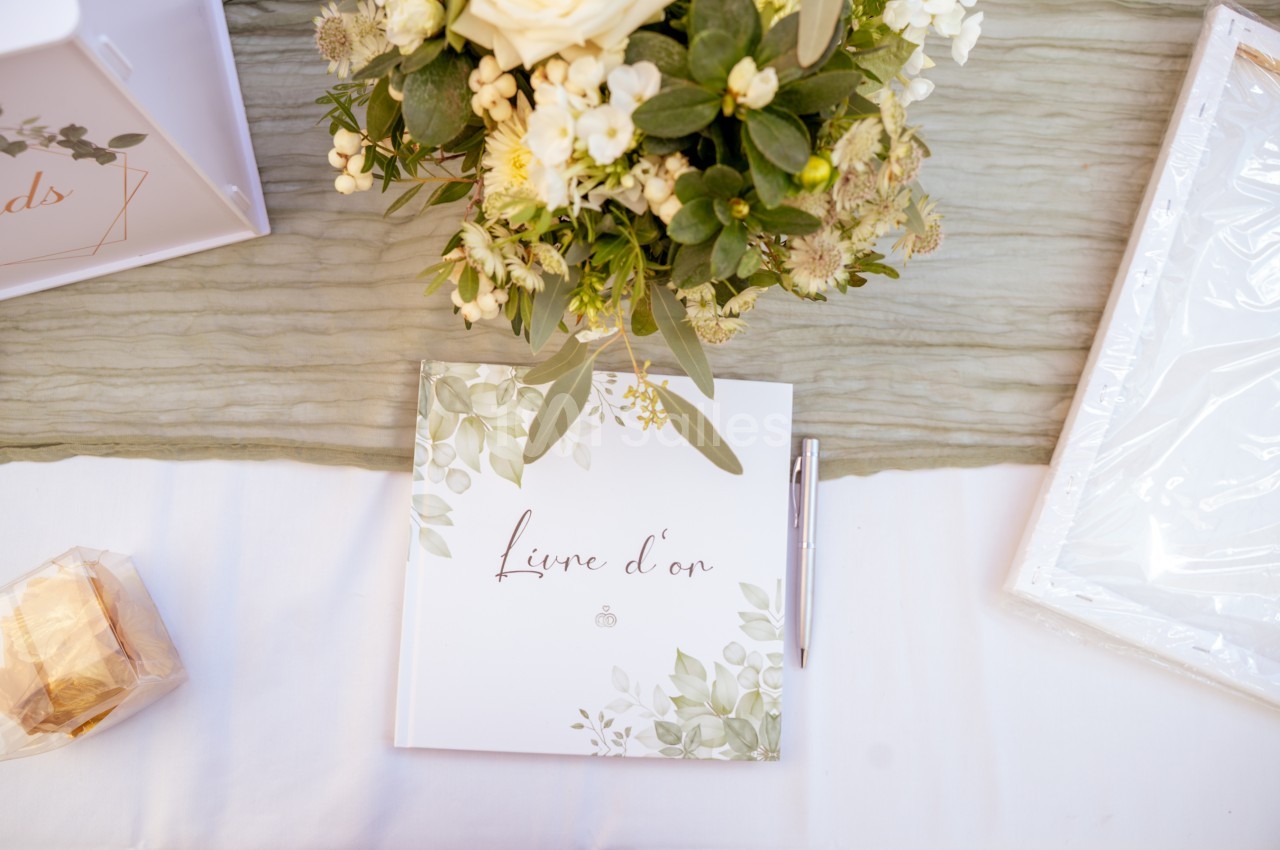 Livre d'or à couverture blanche ornée de feuillages, près d'un bouquet de fleurs crème sur une table blanche.