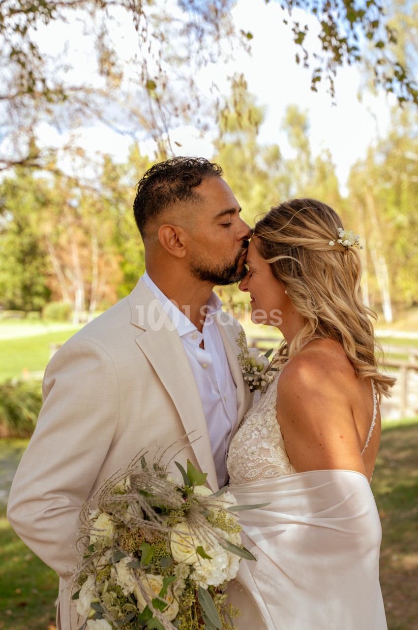 Un homme en costume beige embrasse le front d'une mariée en robe satinée blanche, tenant un bouquet de fleurs blanches, dans un parc arboré en extérieur.