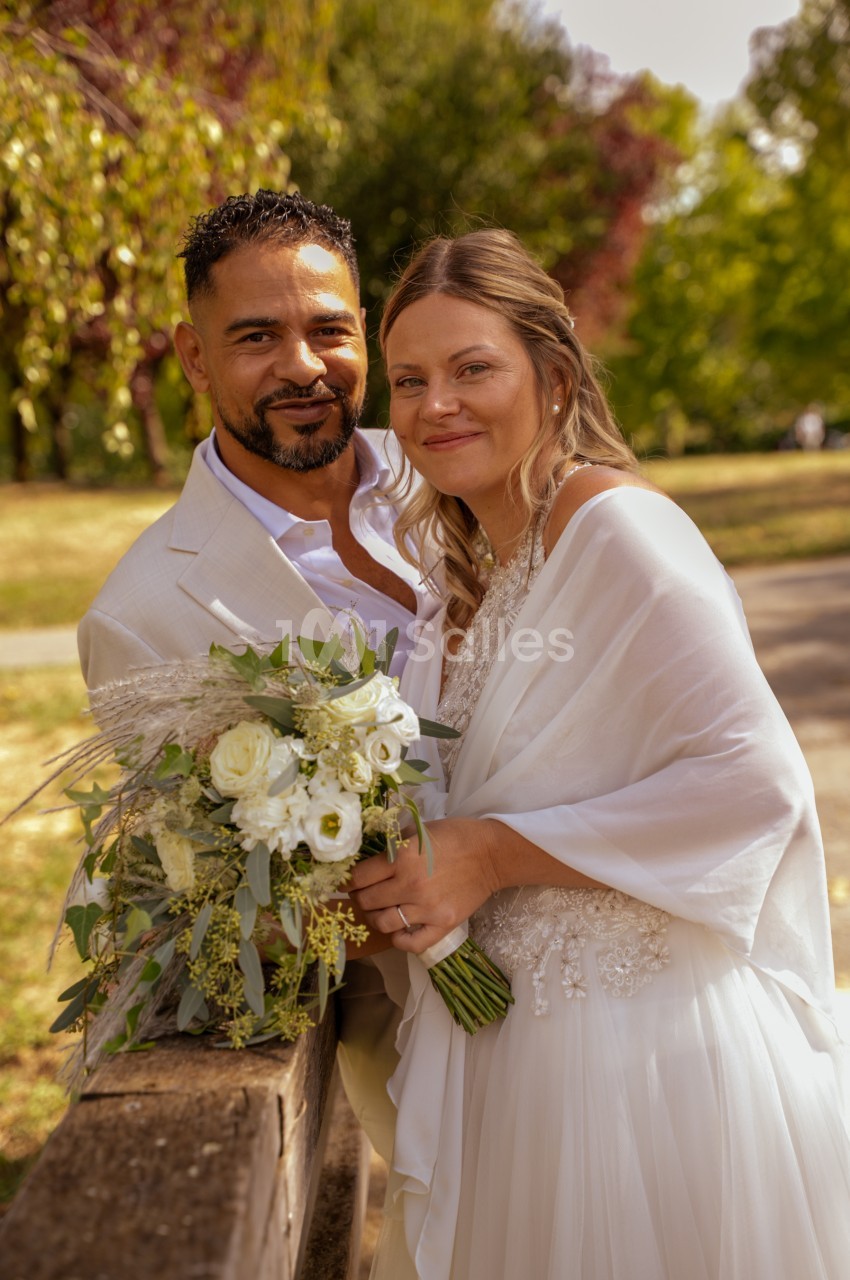 Un couple de mariés souriant, debout en extérieur dans un parc ensoleillé, la mariée tenant un bouquet de roses blanches et d'eucalyptus.