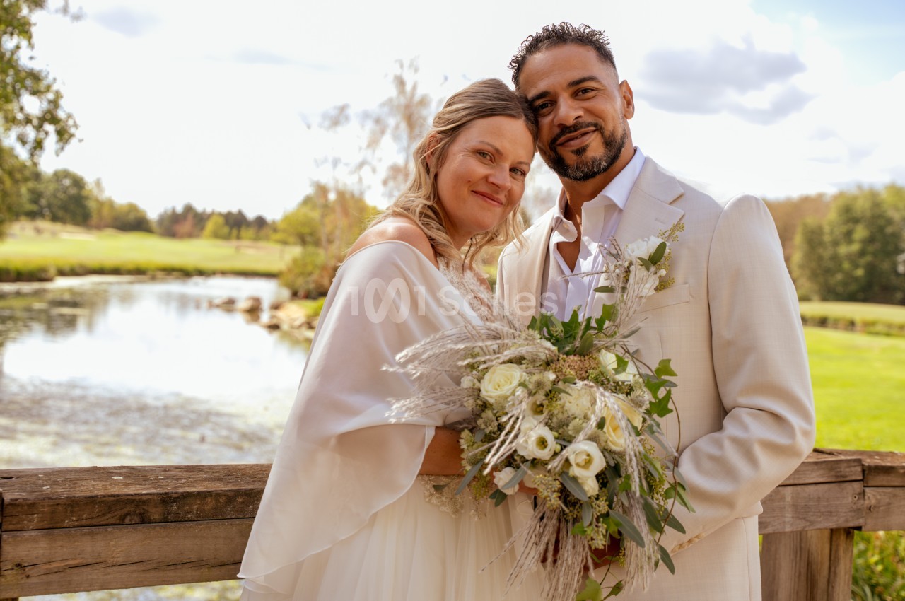Un couple de mariés souriant, debout sur un pont en bois au-dessus d'un plan d'eau, tenant un bouquet de roses blanches et d'herbes sèches.