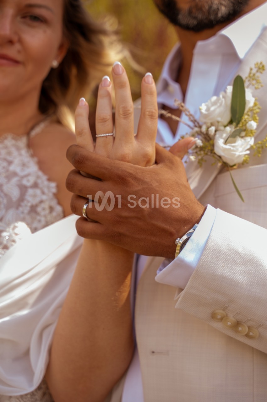 Gros plan sur les mains entrelacées d'une mariée et d'un marié, avec alliances et fleurs blanches au costume.