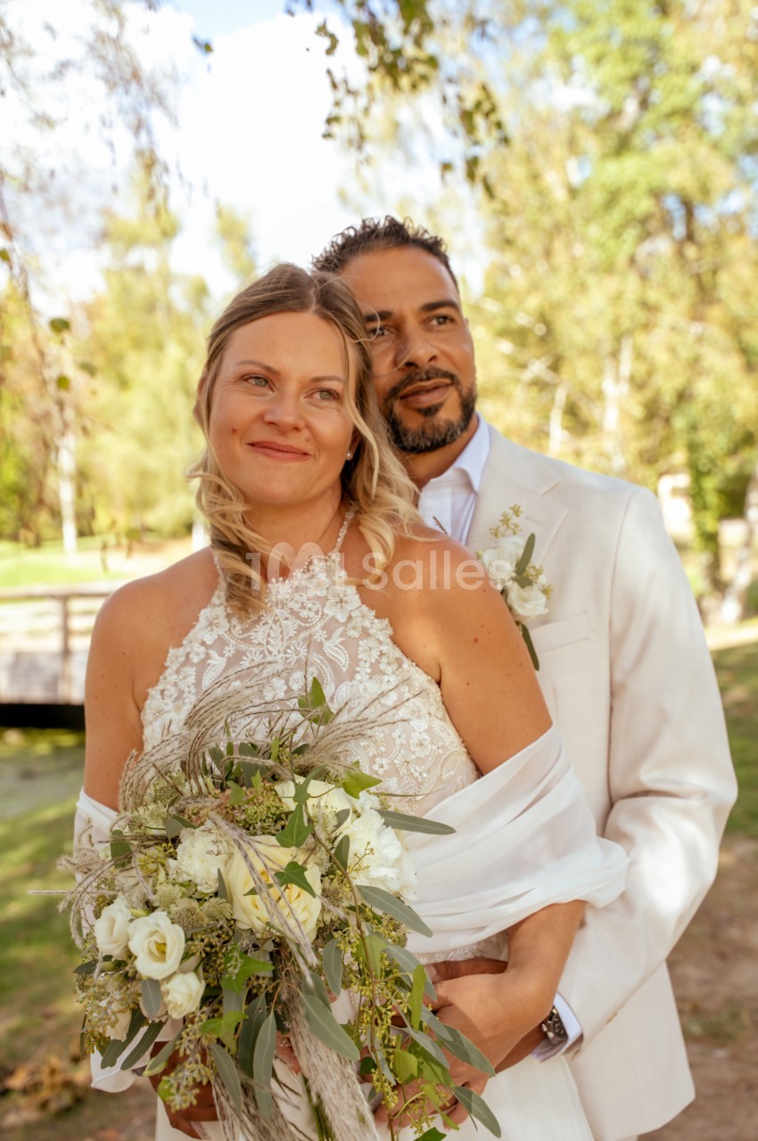 Une mariée au décolleté dentelle et un marié en costume crème, enlacés en extérieur devant des arbres, tenant un bouquet de roses blanches et d'eucalyptus. Let me check length: roughly 165 characters, too long. Need ≤125. Une mariée en robe de dentelle