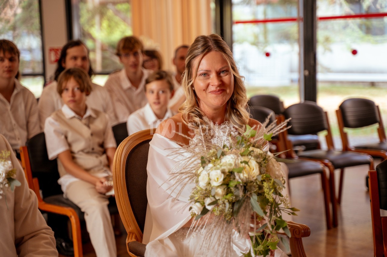 Une mariée blonde en robe blanche tient un bouquet de fleurs blanches et de verdure, assise sur une chaise en bois dans une salle, entourée d'invités attentifs.