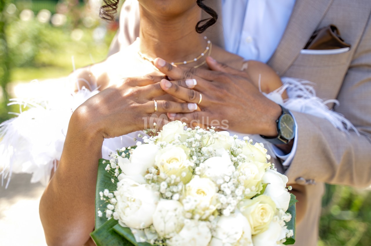 Gros plan sur un bouquet de roses blanches et de gypsophile tenu par une mariée, les mains posées sur la poitrine d'un marié en costume beige. Let me check length: roughly 150 characters, too long. Need ≤125. Shorter: "Bouquet de roses blanches et de