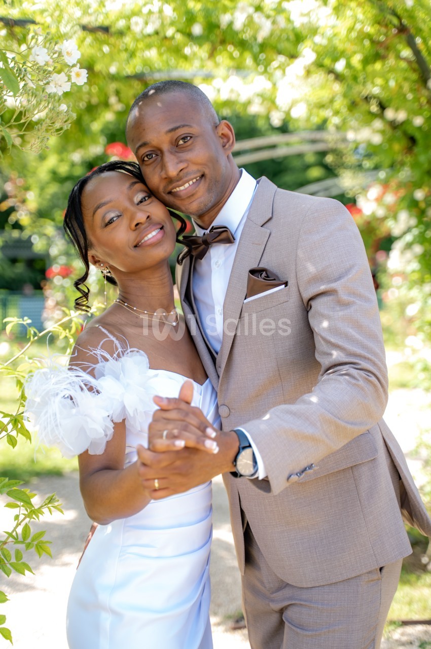 Un couple de mariés souriants, la femme en robe blanche à fleurs et l'homme en costume beige avec nœud papillon marron, pose dans un jardin ombragé de verdure.
