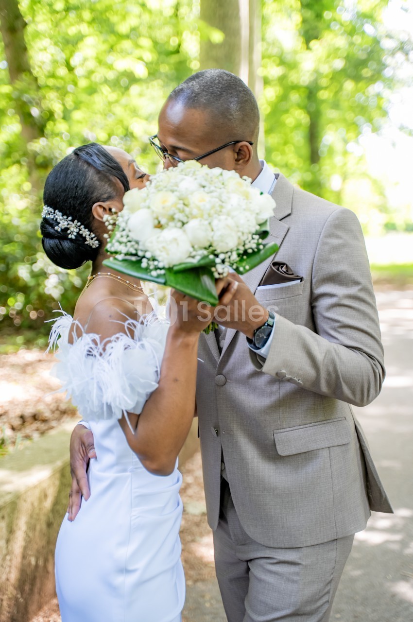 Un couple de mariés s'embrasse derrière un gros bouquet de pivoines blanches en extérieur, sous des arbres verts ; la mariée porte un bijou de cheveux et une robe blanche à plumes, le marié un costume beige. Compte: this is about 215 characters, too