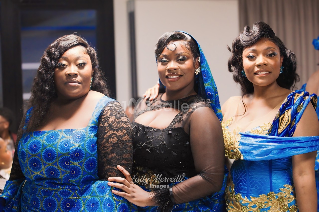 Trois femmes posent côte à côte en robes bleues à motifs wax et dorés, celle du centre portant une tenue noire en dentelle et tulle. Let me check length: about 125 chars? Count roughly: "Trois femmes posent côte à côte en robes bleues à motifs wax et