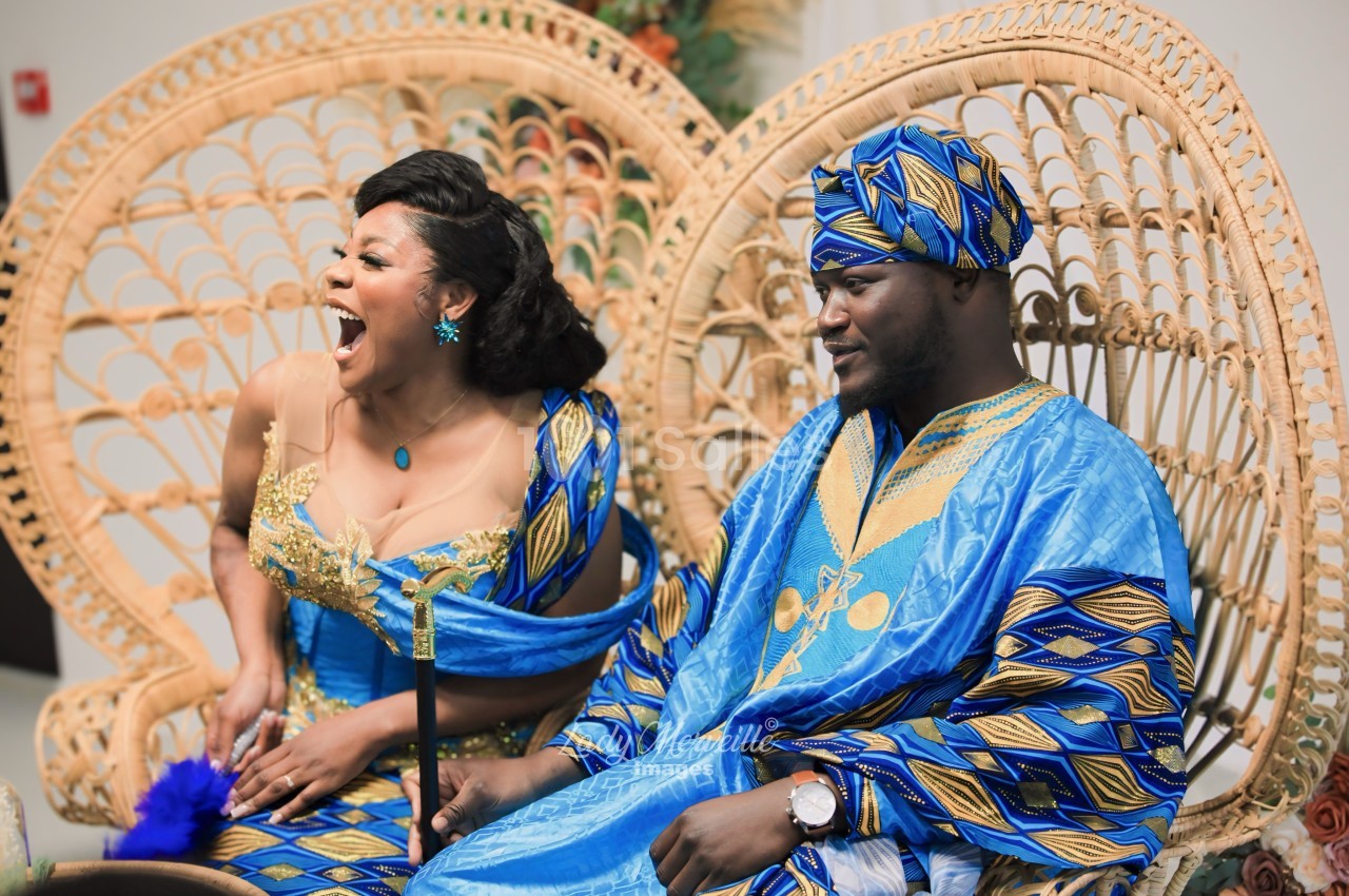 Une femme riant aux éclats et un homme souriant, assis côte à côte dans des fauteuils en rotin, vêtus de tenues bleues et dorées aux motifs wax.