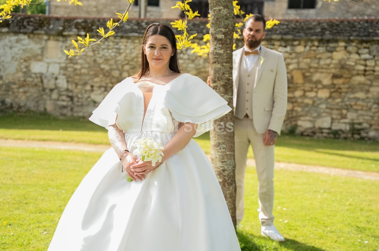 Une mariée en robe blanche volumineuse à larges volants se tient devant un arbre, tandis qu'un marié en costume beige la regarde depuis l'arrière-plan, sur une pelouse devant un mur en pierre.