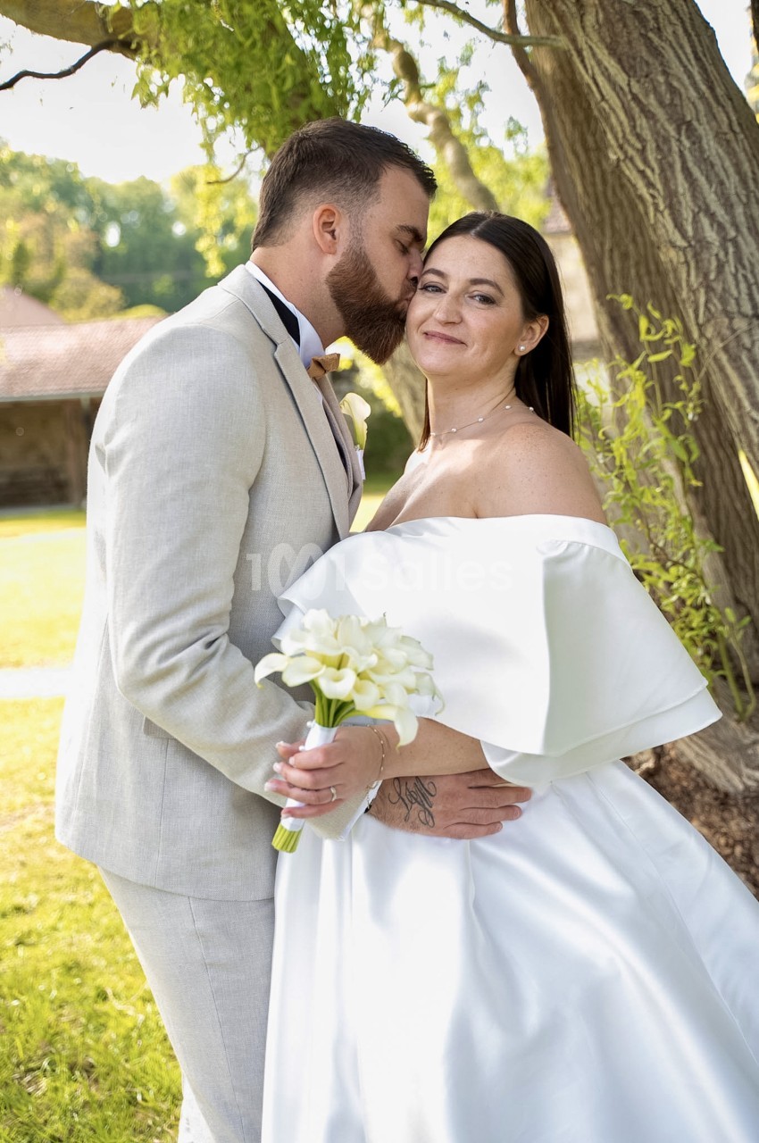 Un homme en costume de lin beige embrasse la joue d'une mariée en robe blanche à larges épaules, qui tient un bouquet de callas, devant un grand arbre en extérieur.