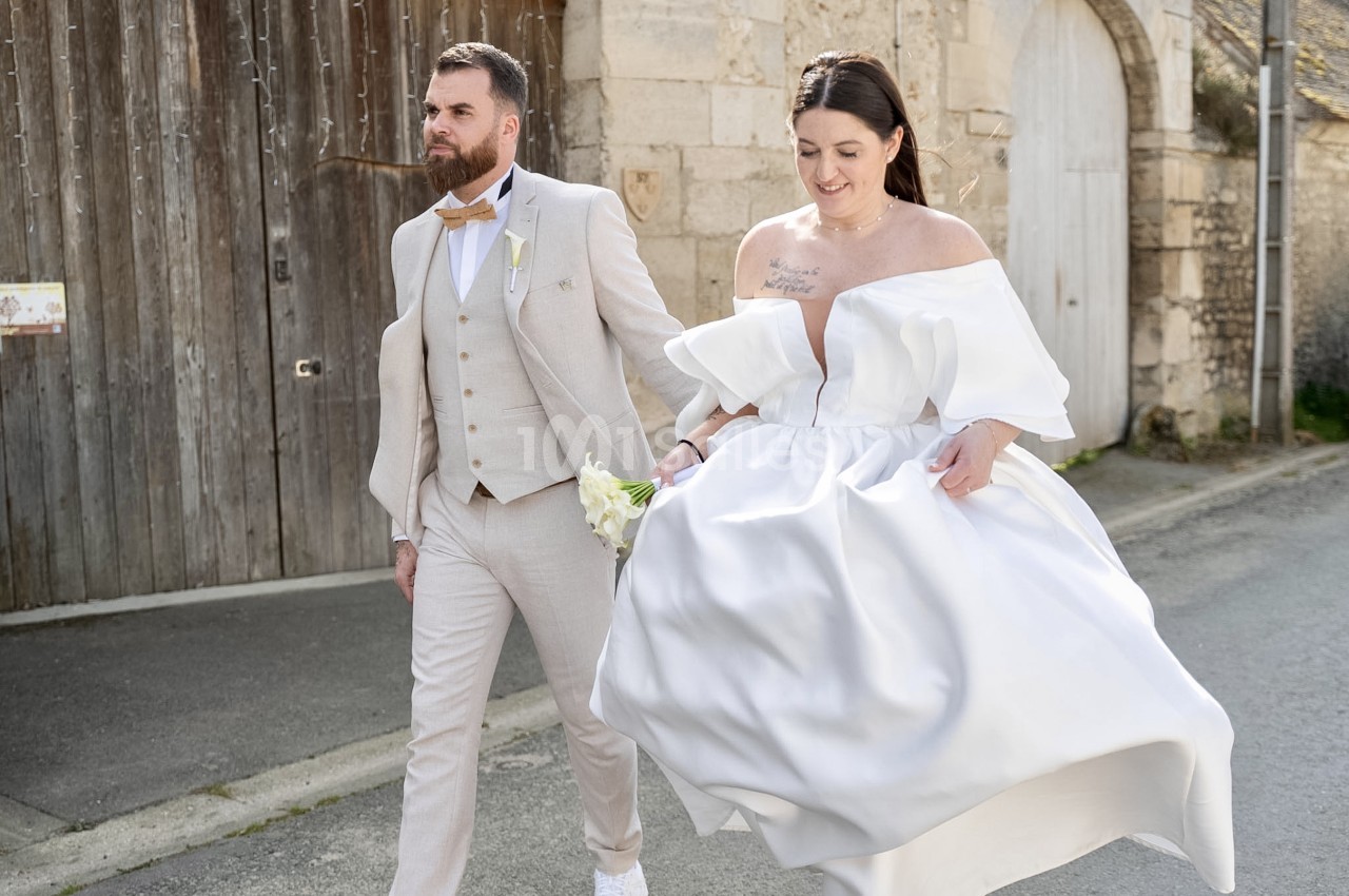 Un couple de mariés marche dans une rue pavée devant un portail en bois et un mur de pierre, la robe blanche de la mariée tourbillonnant au vent. Let me check constraints: under 125 characters? Count roughly: "Un couple de mariés marche dans une rue
