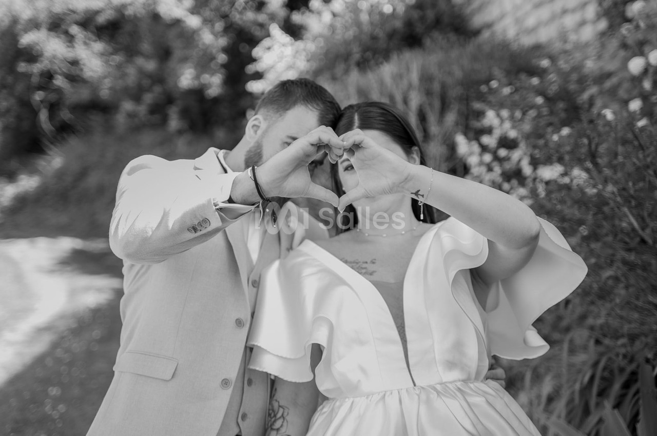Photo en noir et blanc d'un couple en extérieur formant un cœur avec ses mains devant le visage, lui en costume clair, elle en robe blanche à volants, entourés de végétation.