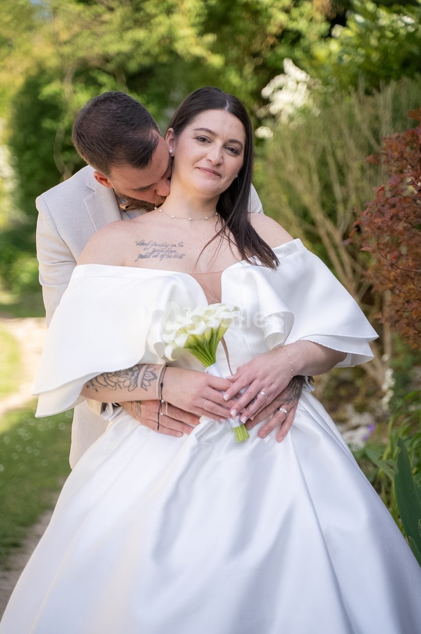 Une mariée en robe blanche à volants tient un bouquet de callas, enlacée par derrière par un marié en veste claire qui l'embrasse dans le cou, dans un jardin verdoyant.