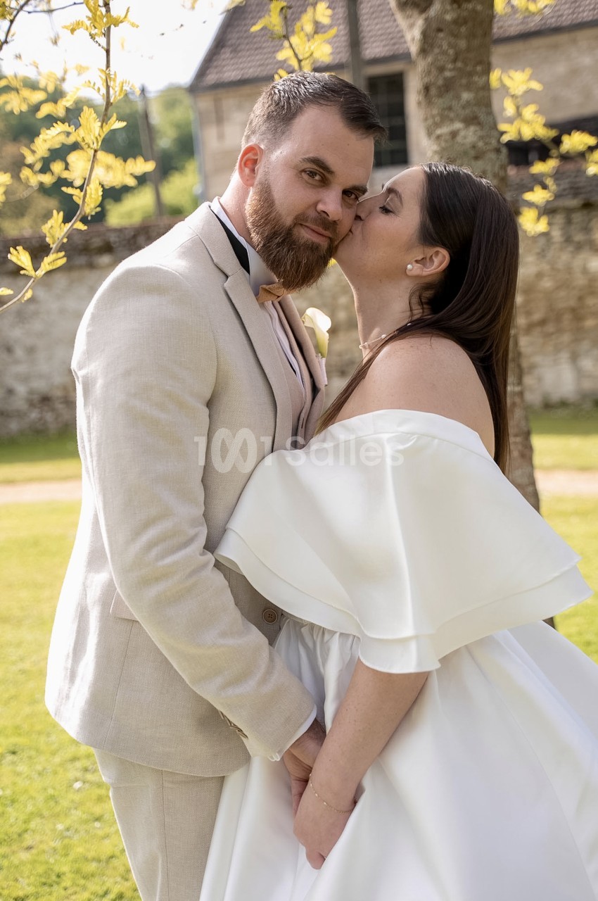 Un couple de mariés en extérieur sur une pelouse, l'homme en costume beige embrasse la joue de la femme en robe blanche à épaules dénudées, près d'un arbre et d'un mur en pierre.