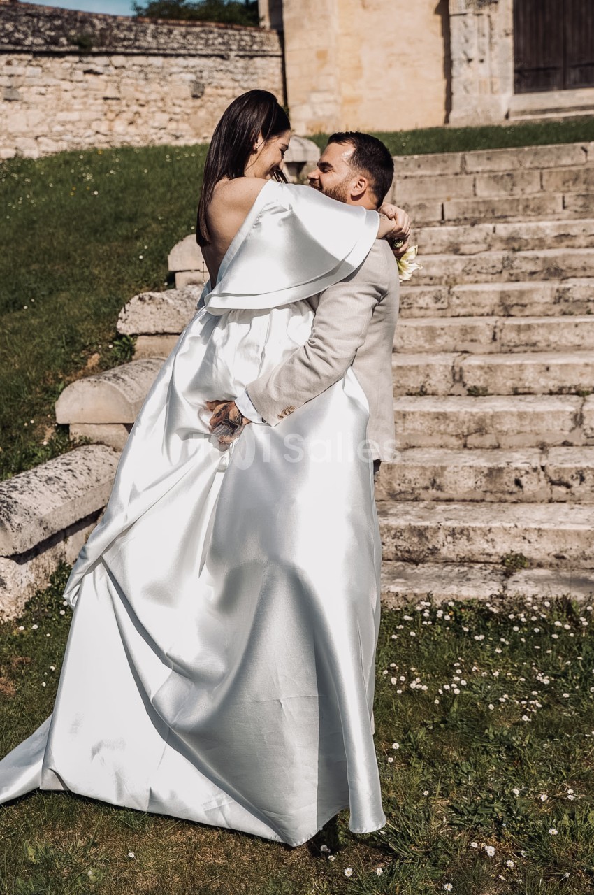 Une mariée en robe de satin blanc à épaule dénudée enlace un marié en costume beige devant un escalier de pierre en extérieur, sur une pelouse.