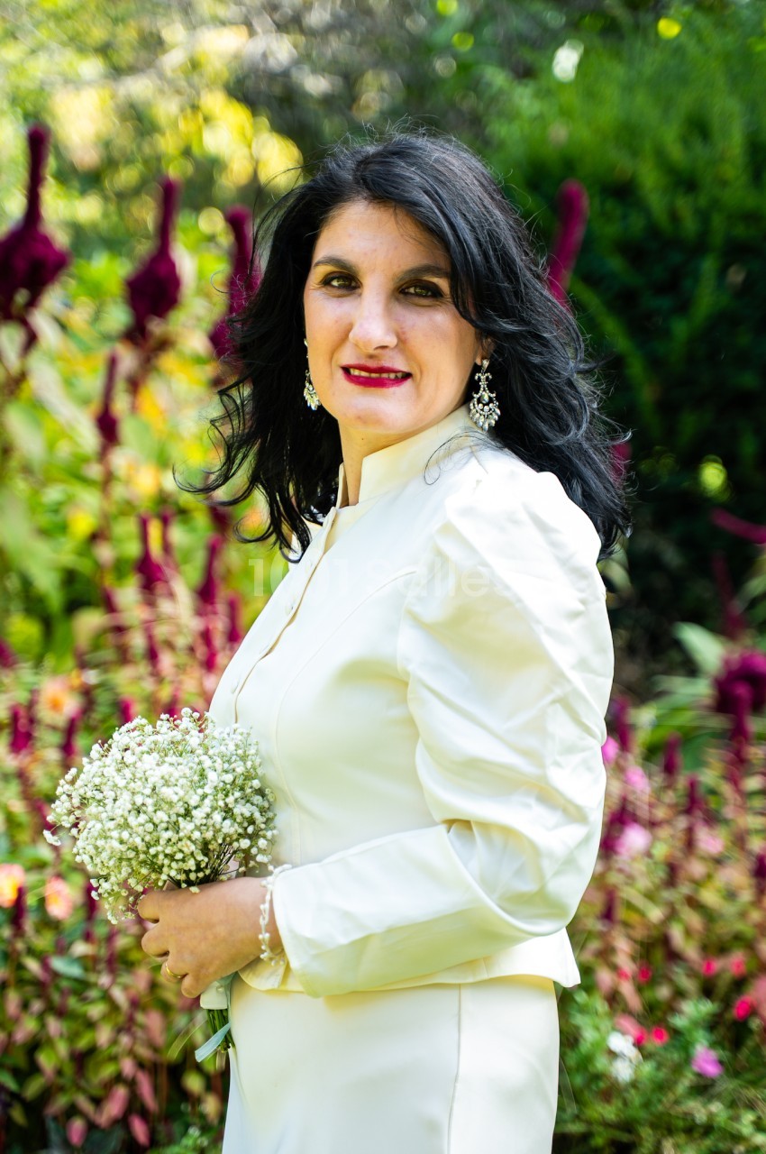 Femme brune souriante, vêtue d'un ensemble ivoire, tenant un bouquet de gypsophile blanc, dans un jardin fleuri aux tons verts et bordeaux.