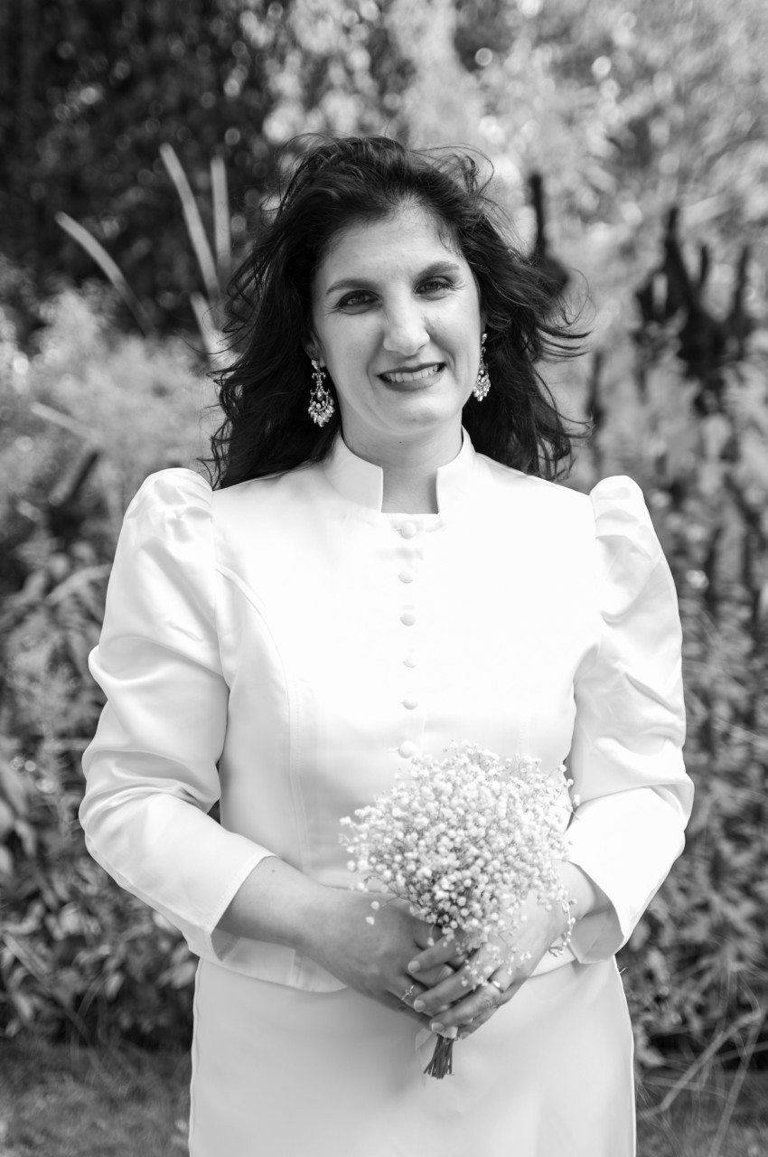 Portrait en noir et blanc d'une femme souriante aux cheveux longs et ondulés, vêtue d'un ensemble blanc à col montant et manches bouffantes, tenant un petit bouquet de gypsophile.
