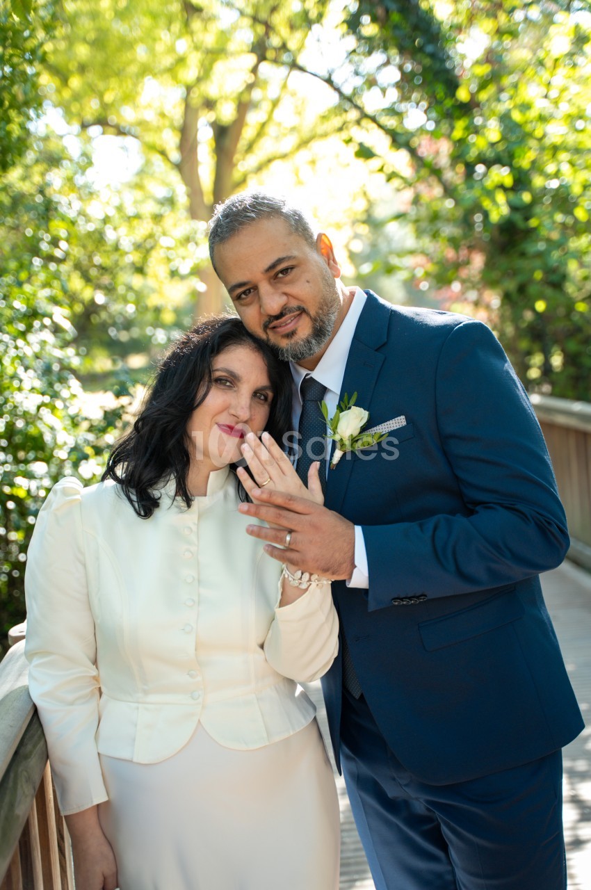 Un couple de mariés pose en extérieur sous des arbres verdoyants, la femme en veste crème tenant la main de l'homme en costume bleu marine orné d'une fleur blanche. Vérification : environ 170 caractères, trop long. Reformulation : Un couple de mariés