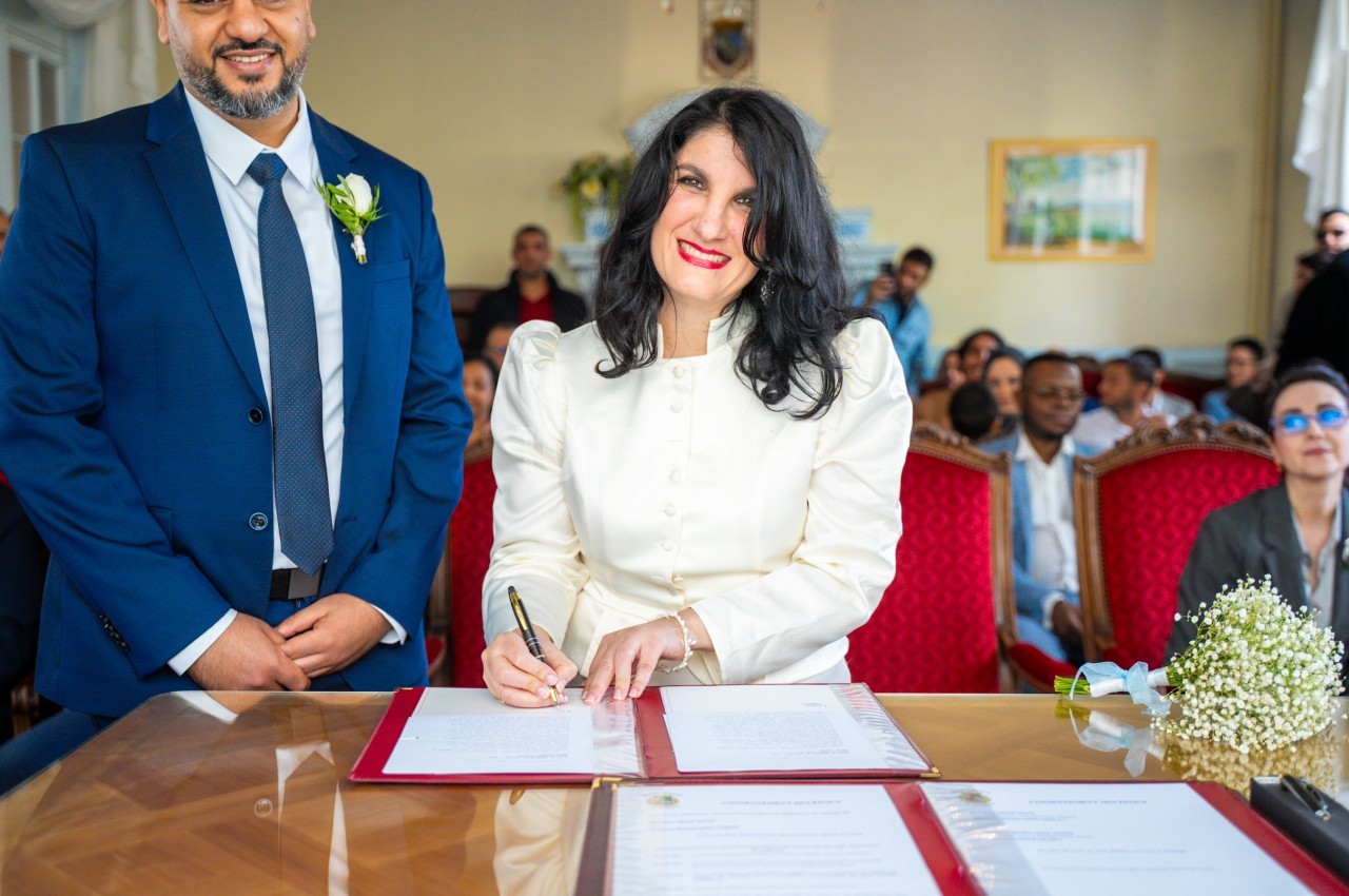 Une mariée en tailleur blanc signe un registre sur une table en bois, tandis que le marié en costume bleu se tient à ses côtés et que des invités sont assis derrière eux.