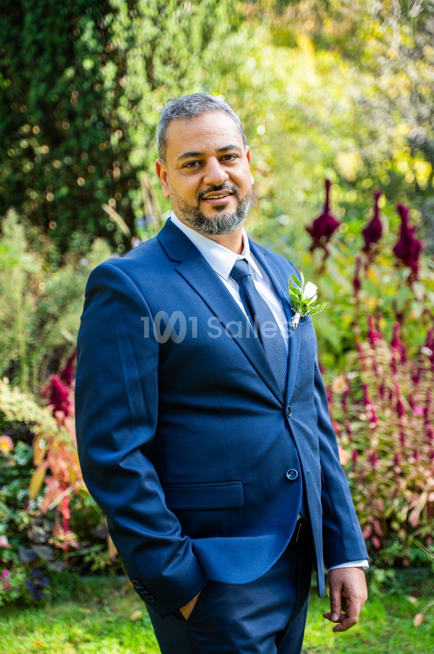 Homme en costume bleu marine, chemise blanche et cravate, avec une fleur à la boutonnière, posant devant un jardin fleuri.