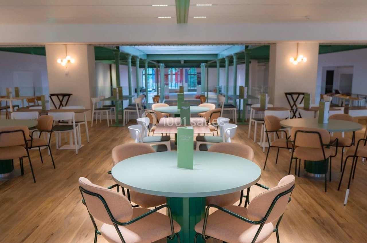 Salle lumineuse avec des tables rondes vertes, des chaises beige et un sol en bois, dans un espace moderne et épuré.