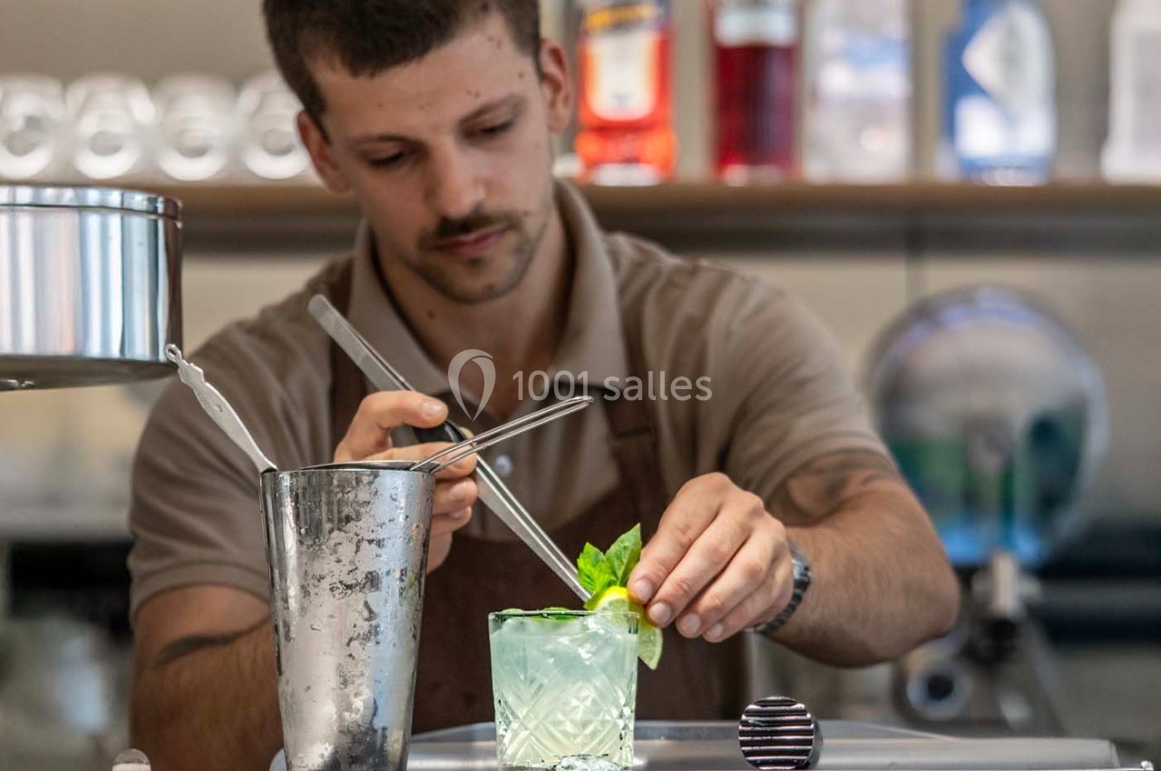 Un barman prépare un cocktail avec des herbes fraîches et une tranche de citron dans un verre transparent.