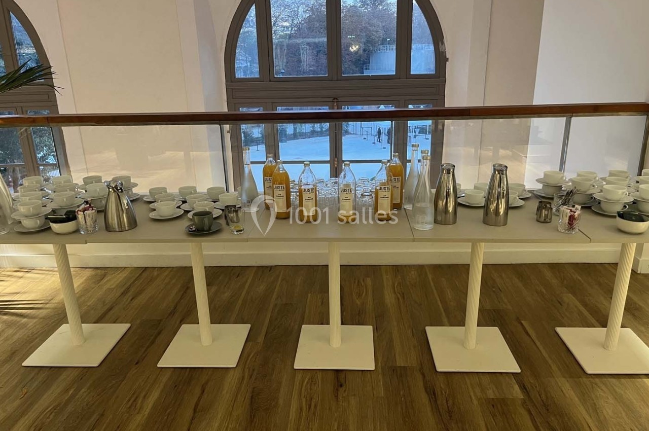 Table dressée avec des carafes de jus, thermos, tasses et bols, placée devant une grande fenêtre cintrée.