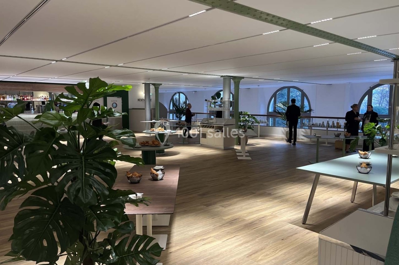 Espace intérieur lumineux avec des plantes, des tables et des personnes debout près de grandes fenêtres arrondies.
