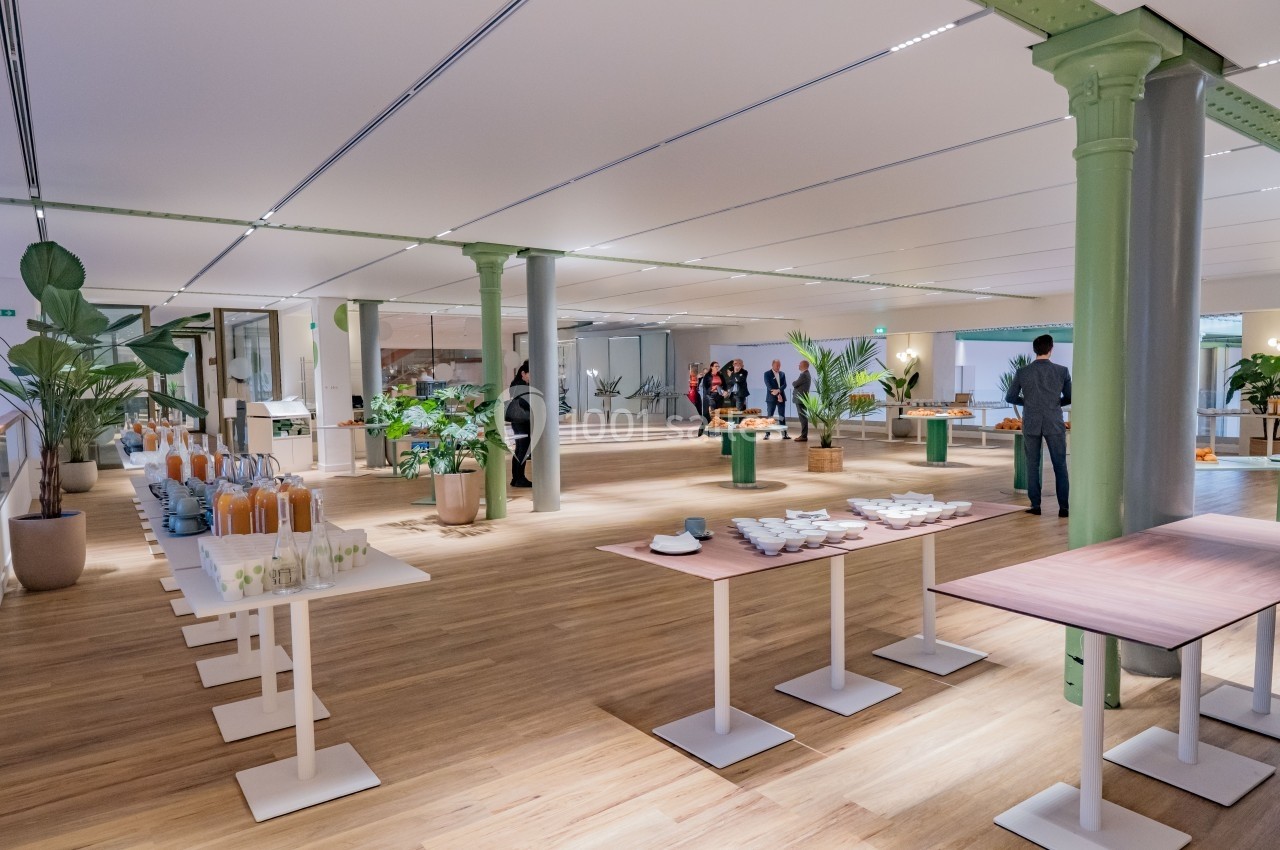 Salle lumineuse avec parquet, tables dressées pour un buffet, plantes décoratives et quelques personnes en arrière-plan.