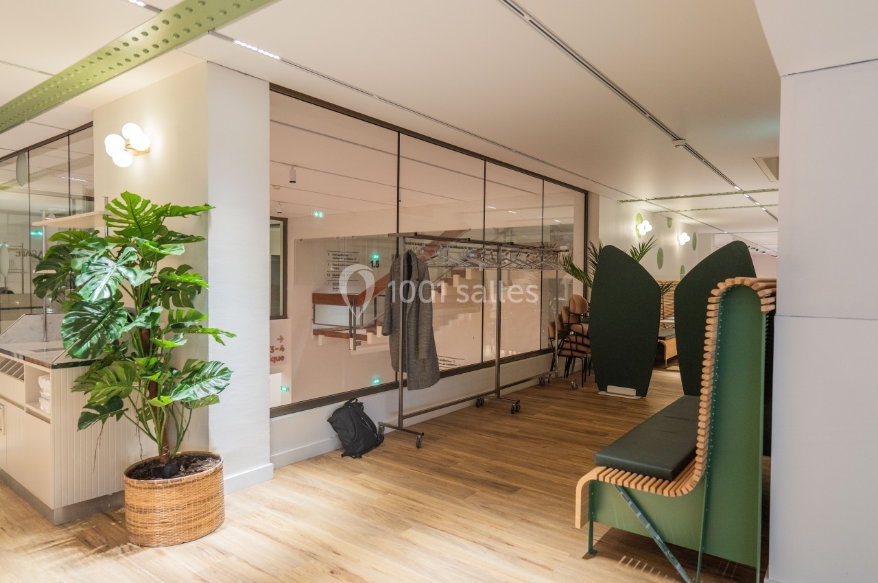 Espace intérieur lumineux avec plantes, mobilier en bois et assises vertes, séparé par une cloison vitrée.