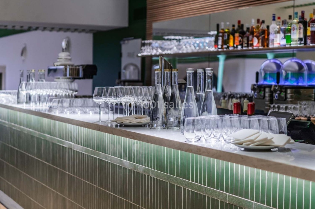 Comptoir de bar moderne avec verres, bouteilles d'eau, assiettes et étagères garnies de bouteilles en arrière-plan.