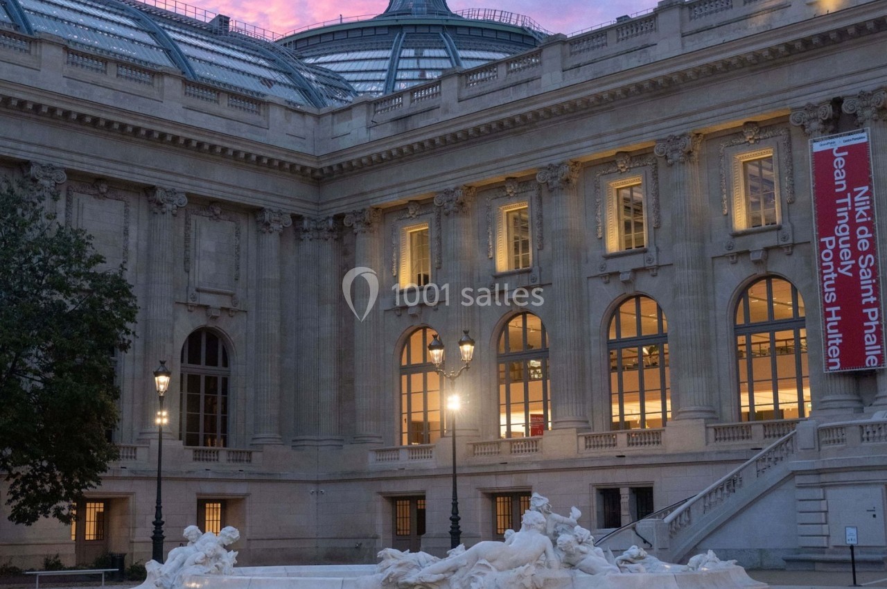 Façade illuminée d'un bâtiment historique avec une verrière, une bannière rouge et une statue au premier plan.