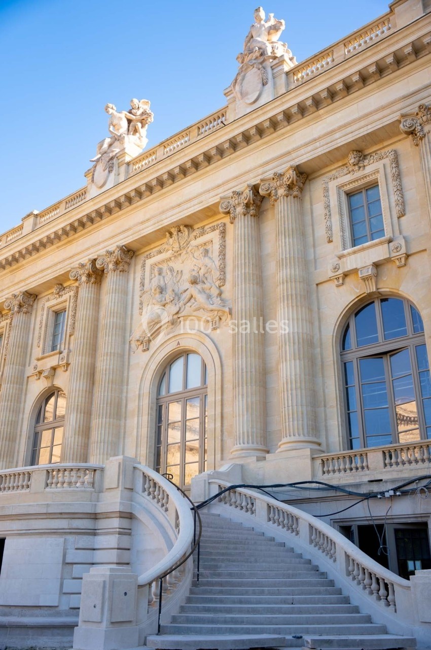 Façade d'un bâtiment historique en pierre avec colonnes, sculptures et escalier courbé sous un ciel bleu.