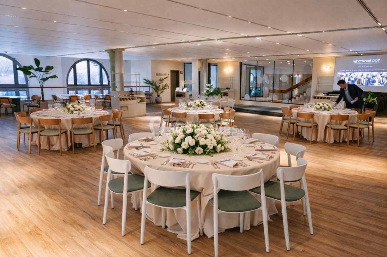 Salle de réception lumineuse avec tables rondes dressées, décorées de bouquets de fleurs blanches et chaises modernes.