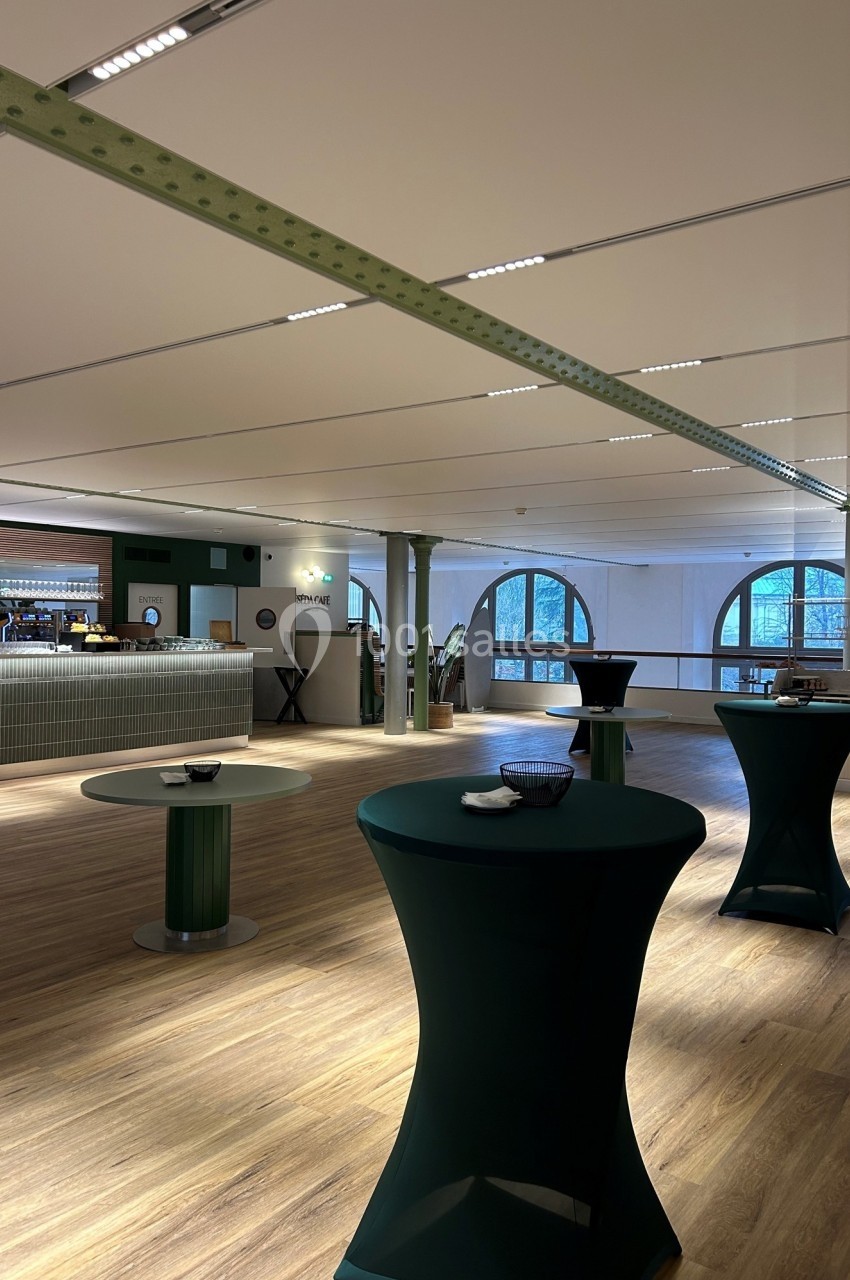 Salle lumineuse avec parquet, tables hautes couvertes de nappes vertes et un bar en arrière-plan.