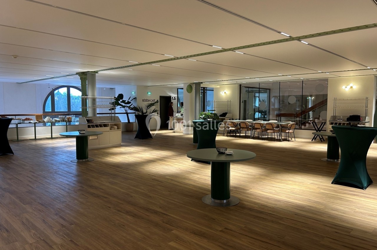 Salle spacieuse avec parquet, tables hautes habillées de nappes vertes et grandes fenêtres laissant entrer la lumière…