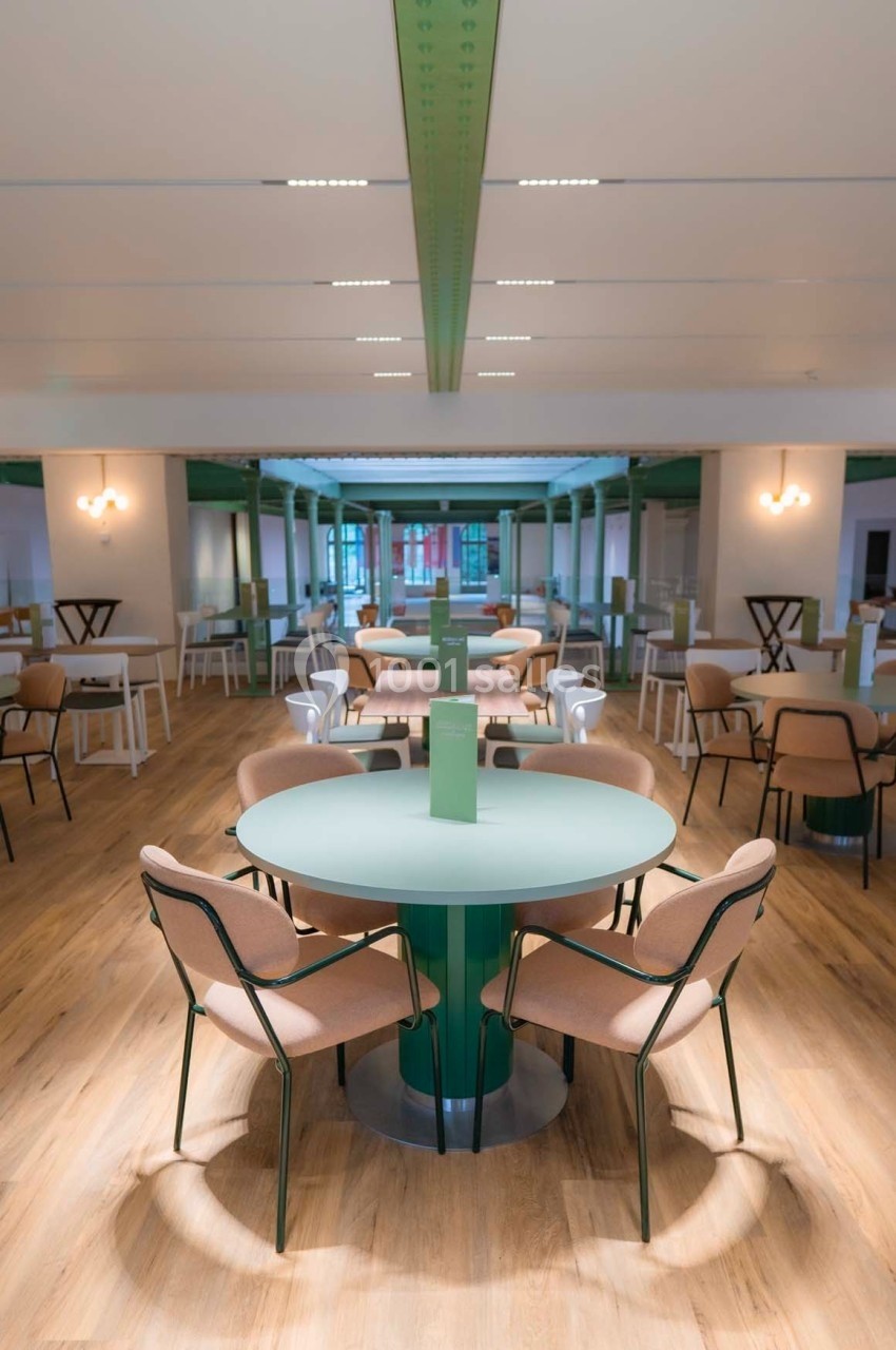 Salle lumineuse avec tables rondes vertes, chaises beige et vert, sol en bois clair et plafond blanc avec poutres vertes.