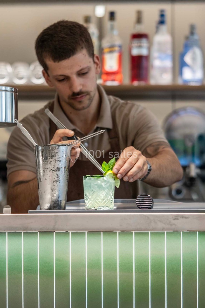Un barman prépare un cocktail vert garni de feuilles de menthe sur un comptoir lumineux.