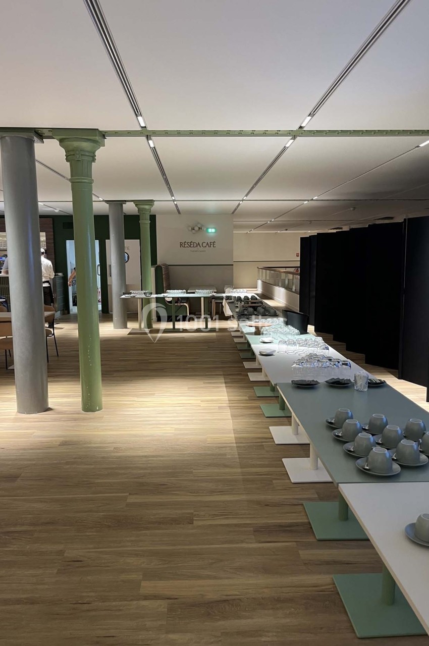 Salle lumineuse avec tables alignées, vaisselle disposée et piliers métalliques, dans un espace moderne et épuré.