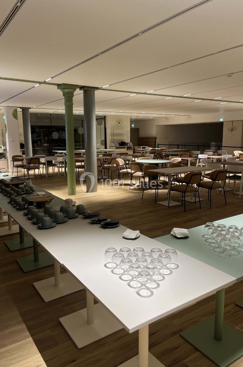 Salle lumineuse avec tables alignées, chaises en bois, et buffets préparés avec verres et vaisselle.