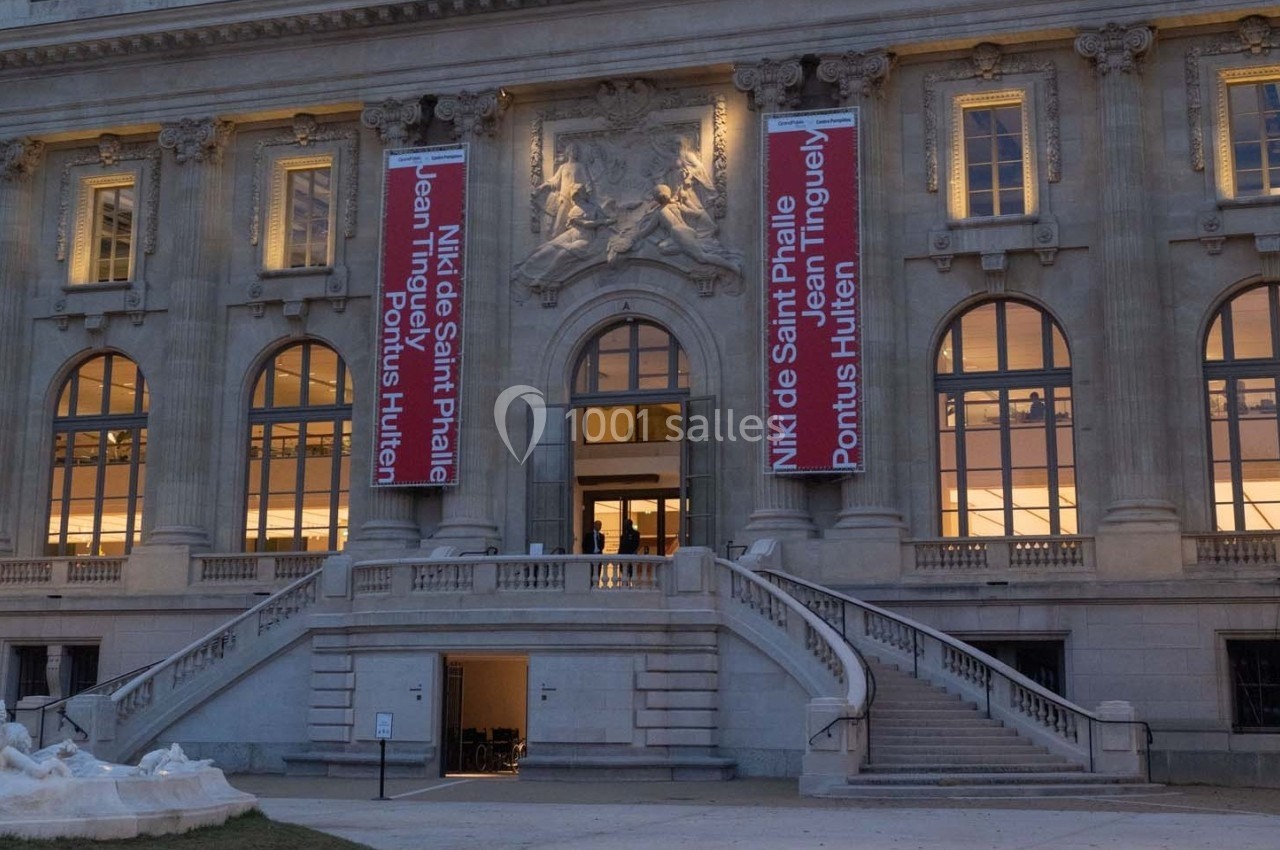 Façade d'un bâtiment historique avec des bannières rouges annonçant une exposition, vue en soirée.