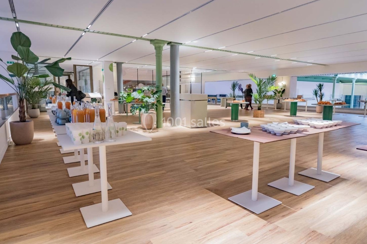 Salle lumineuse avec parquet, tables dressées pour un buffet, plantes décoratives et grandes baies vitrées.